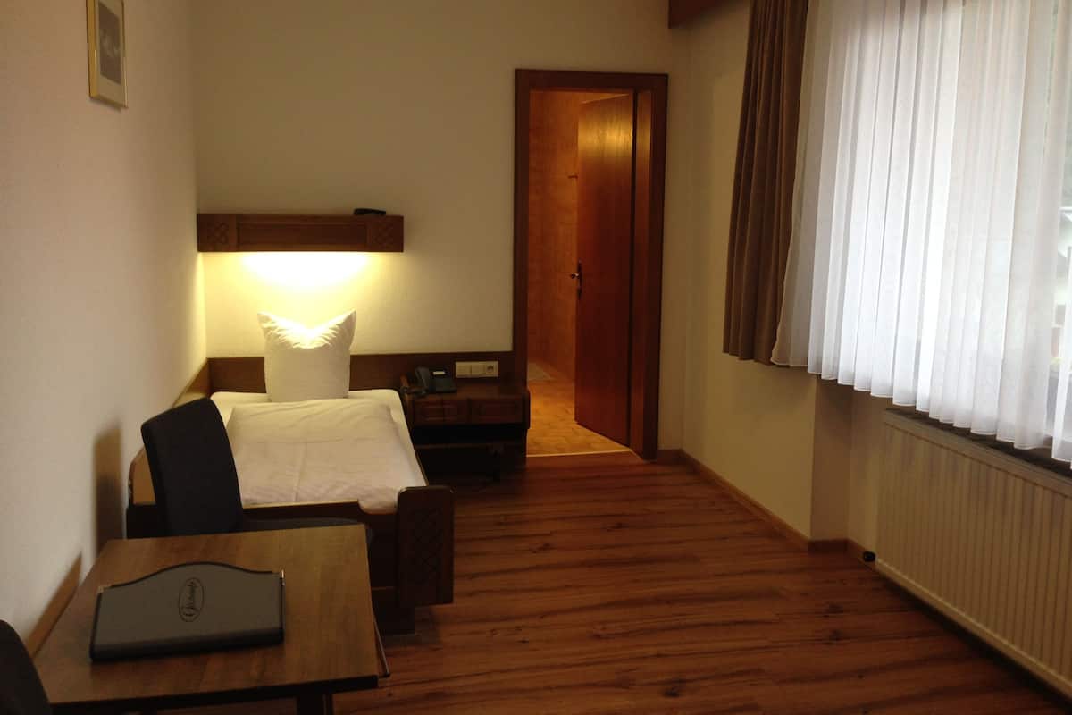 A hotel room with a wooden floor, a desk with a lamp, a bed with white bedding, a nightstand, and a door leading to another room.