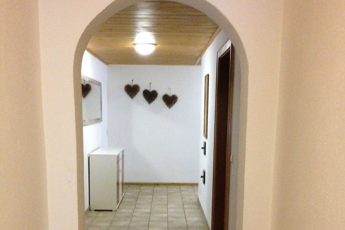 A hallway with a tiled floor, a wooden ceiling, and a white door at the end. There are three heart-shaped decorations on the wall.
