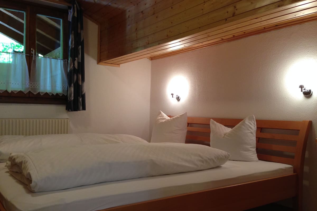 A single bed with white bedding in a room with wooden beams on the ceiling and a window with curtains.