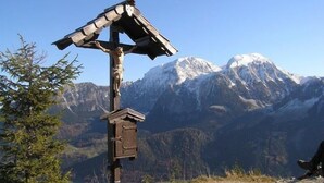 Une croix en bois, sur laquelle est fixée une petite nichée à oiseaux, se dressant sur fond de montagnes enneigées.
