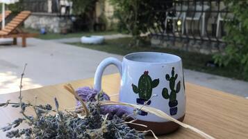 Una mesa con una taza blanca decorada con diseños de cactus, una pequeña planta en maceta y un banco de madera en un patio con flores rosadas y un edificio al fondo.
