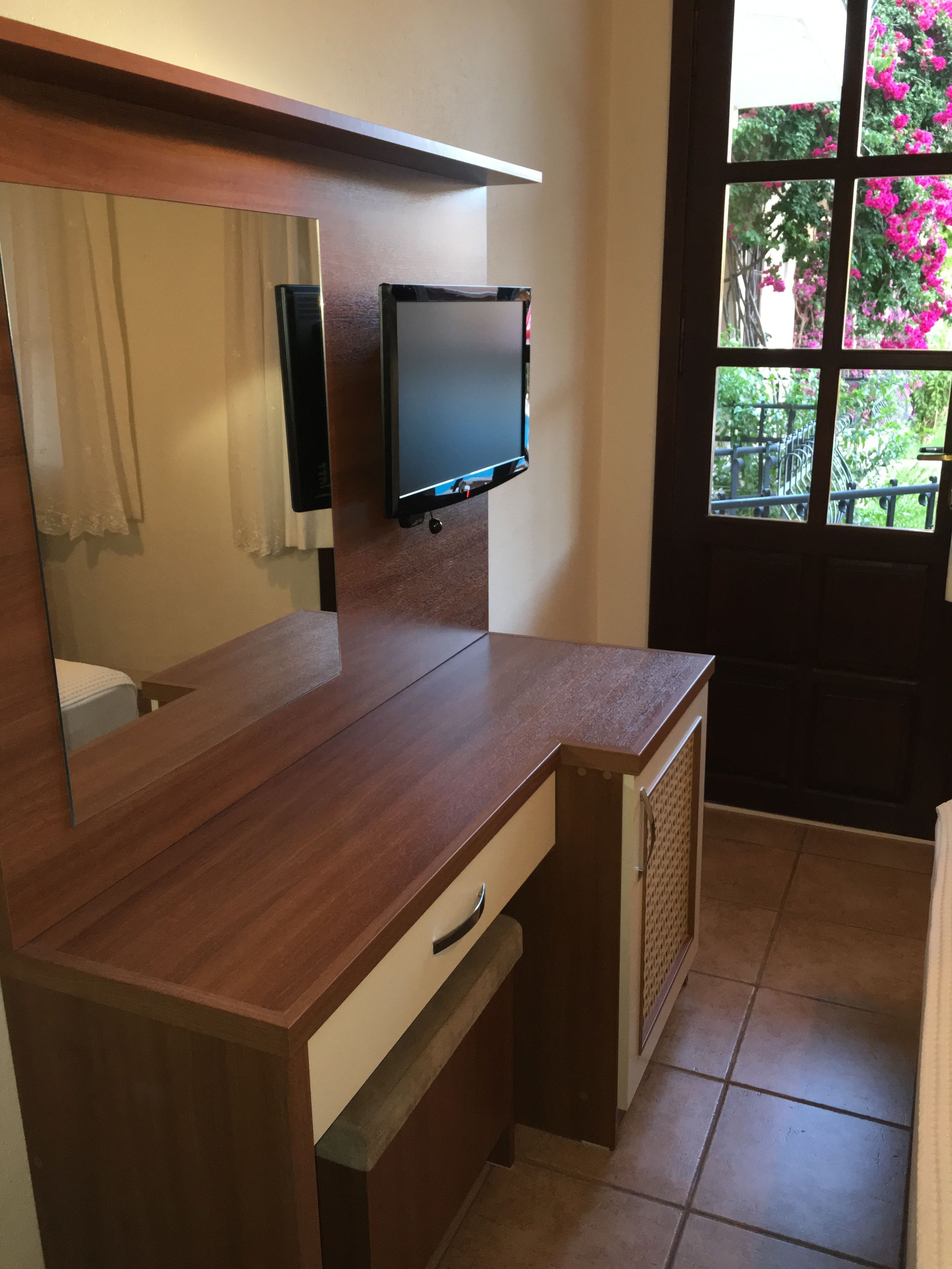 A wooden desk with a television mounted above it, a mirror, and a window with a view of greenery.