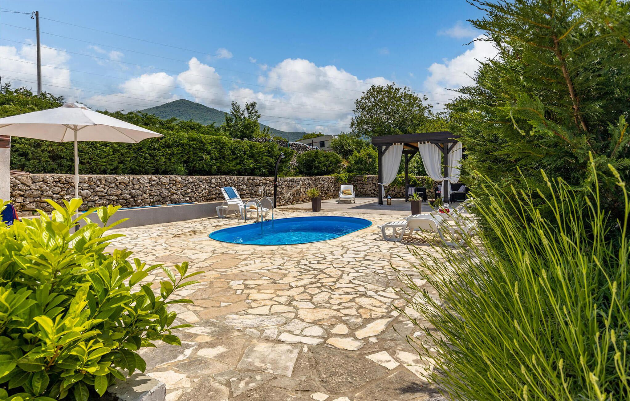A stone-paved outdoor area with a swimming pool, lounge chairs, and a pergola with white curtains.