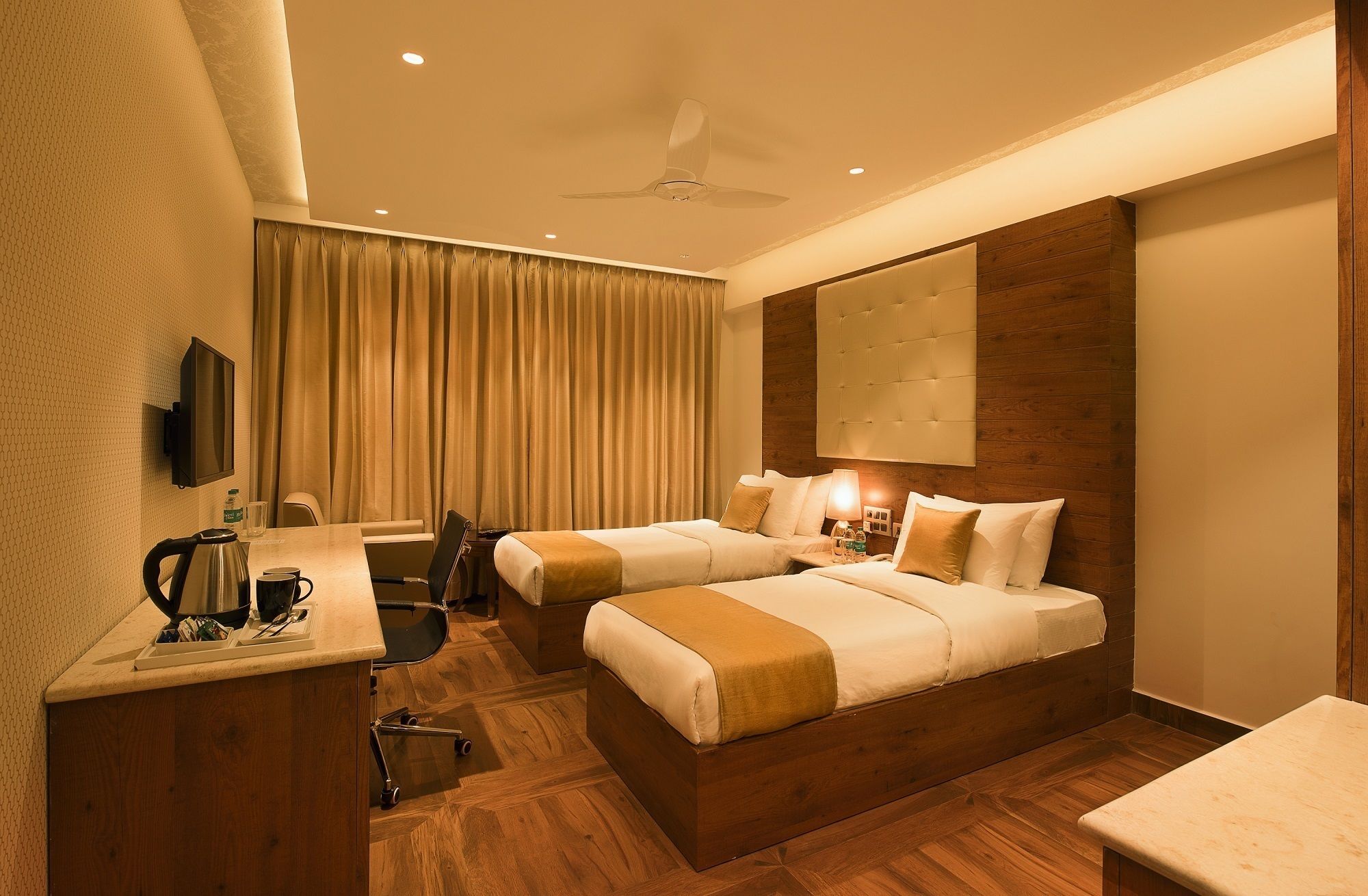 A hotel room with two beds, a desk with a kettle, a chair, a TV, and a ceiling fan.