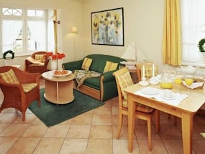 Una sala de estar acogedora con un sofá verde, sillas de mimbre, una mesa de comedor lista para el desayuno y un cuadro en la pared.
