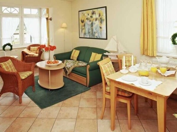 Un salon chaleureux avec un canapé vert, des fauteuils en osier, une table à manger dressée pour le petit-déjeuner et un tableau accroché au mur.