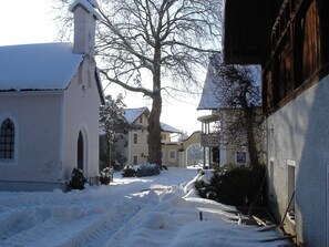 Rua coberta de neve com edifícios, incluindo uma igreja com uma torre e uma casa amarela com varanda.