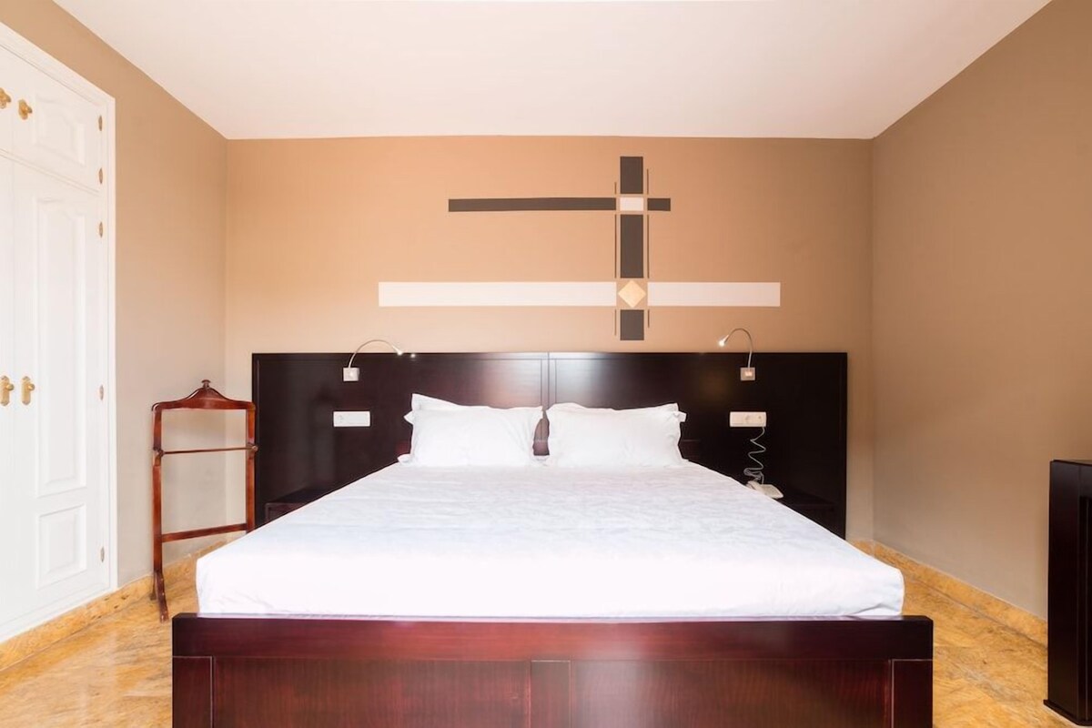 Una cama bien hecha en una habitación con una cruz grande en la pared, un cabecero de madera y un taburete de madera pequeño.
