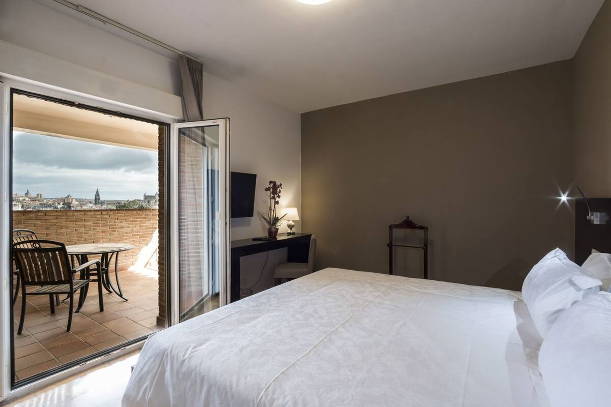 A hotel room with a bed, a desk, a chair, and a view of the city through a glass door.