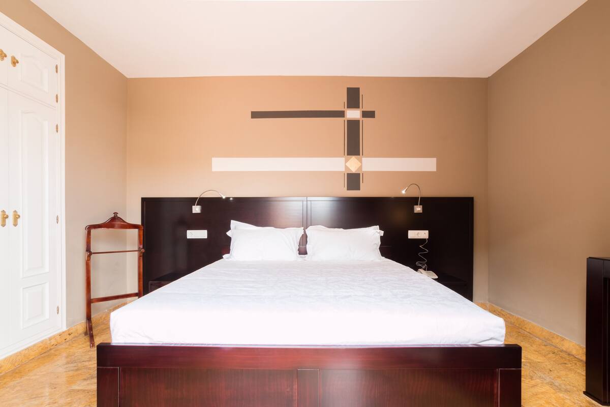 A neatly made bed in a room with a large cross on the wall, a wooden headboard, and a small wooden side table.