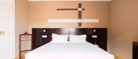 Una cama bien hecha en una habitación con una gran cruz en la pared, un cabecero de madera y una mesita de noche de madera.