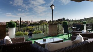 Una terraza en la azotea con mobiliario de mimbre, una mesa con cubierta de vidrio y vistas a un paisaje urbano de fondo.