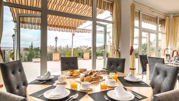 Una mesa de comedor redonda con sillas, alimentos para el desayuno y bebidas en una habitación luminosa con ventanas amplias y vistas a la ciudad.