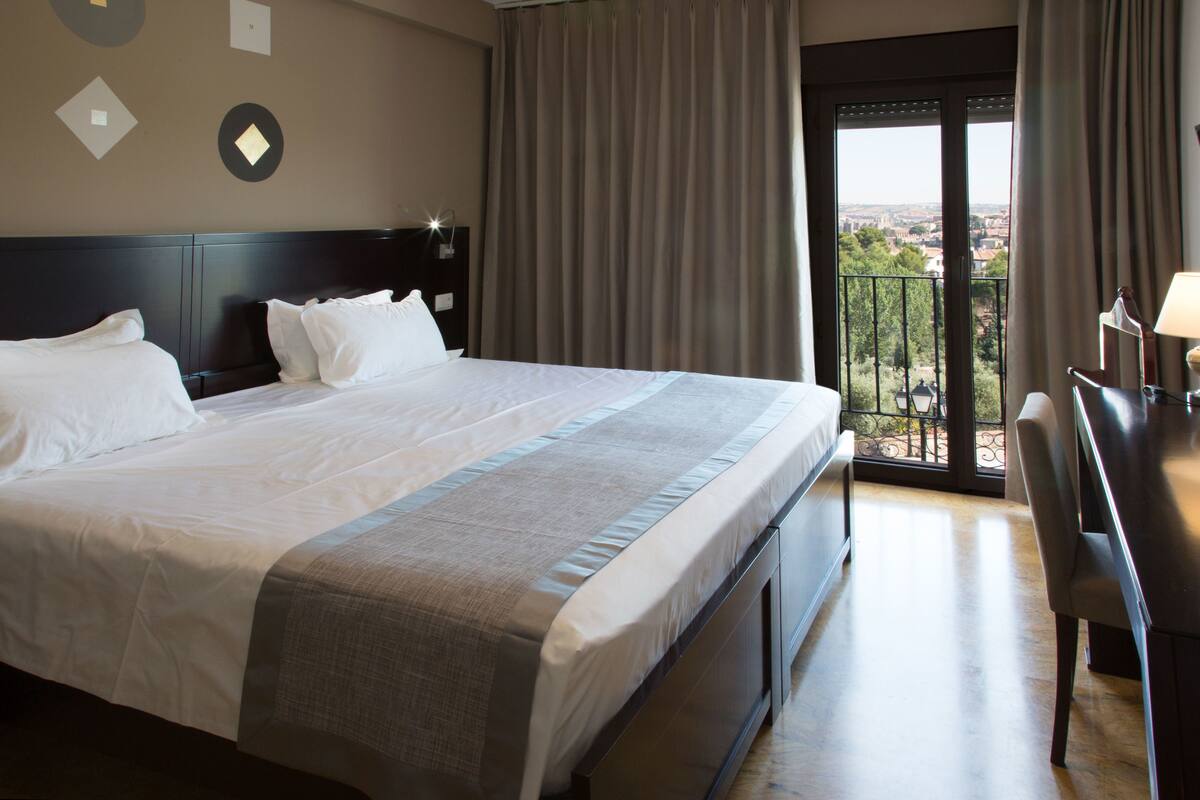 A hotel room with a large bed, a desk with a chair, and a view of the city through the window.