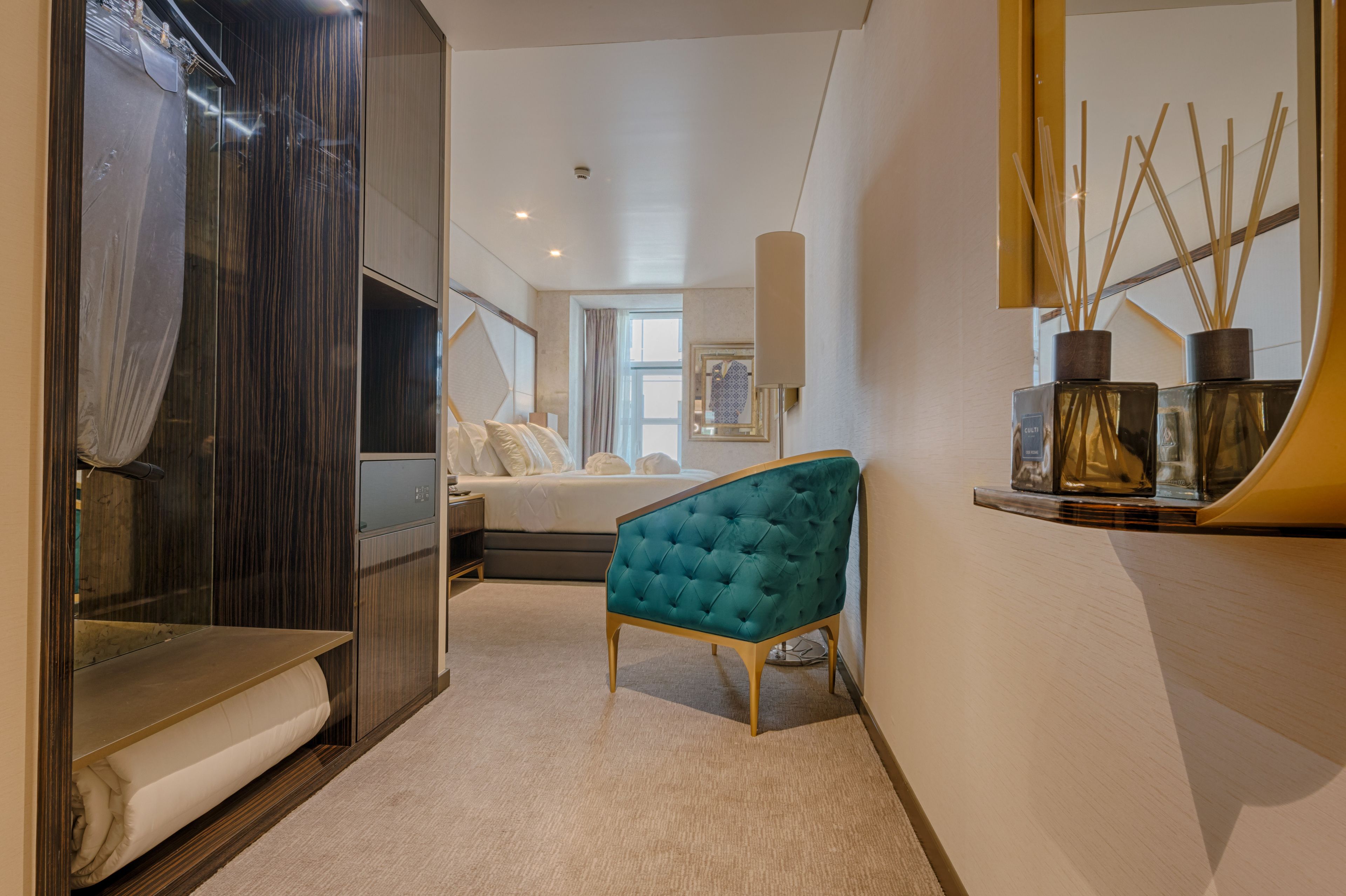 Una habitación de hotel moderna con una cama grande, una zona de estar con un sillón azul, un tocador con espejo y un estante de pared con adornos.