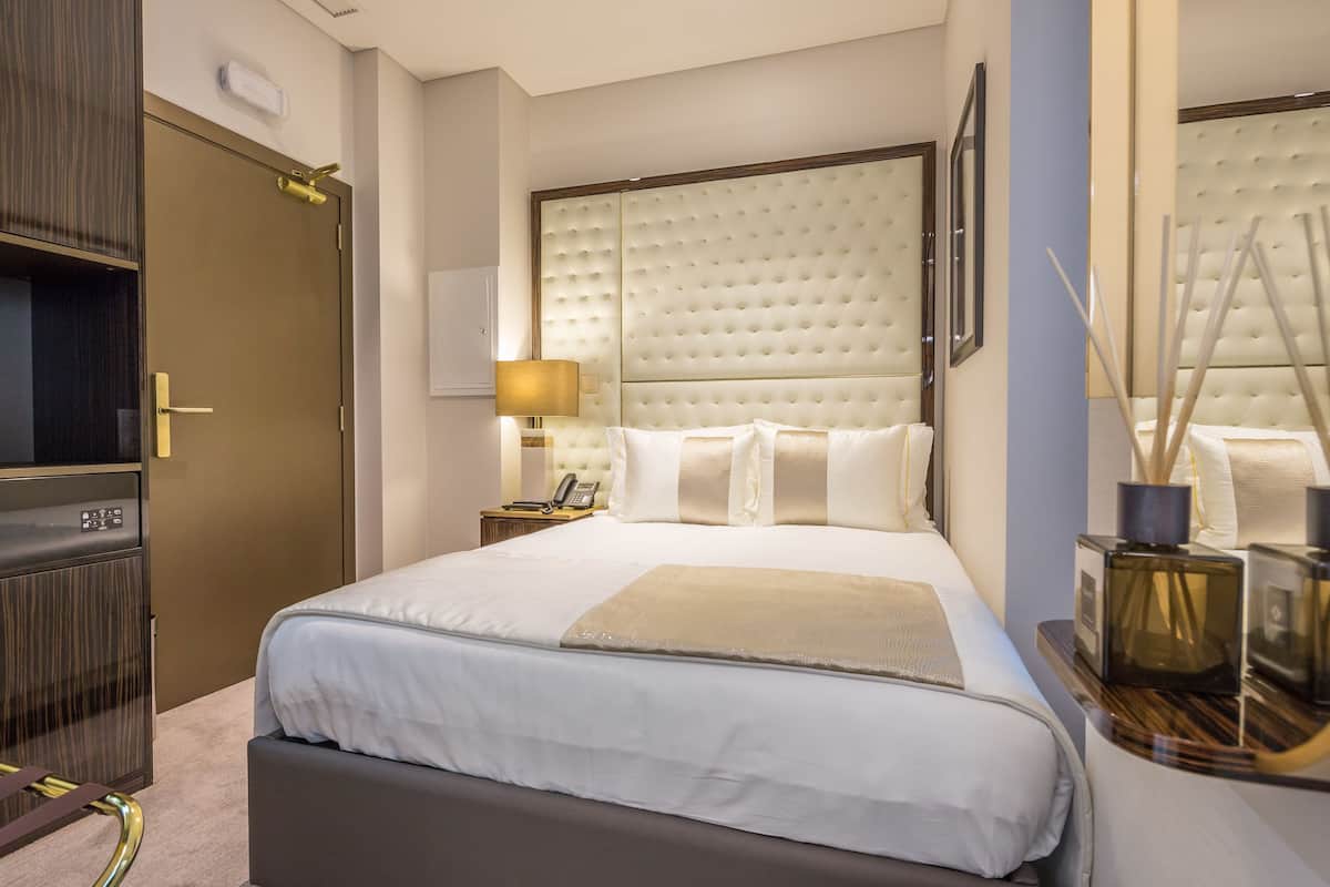 A hotel room with a large bed, a bedside table with a lamp, a minibar, and a door with a keypad lock.