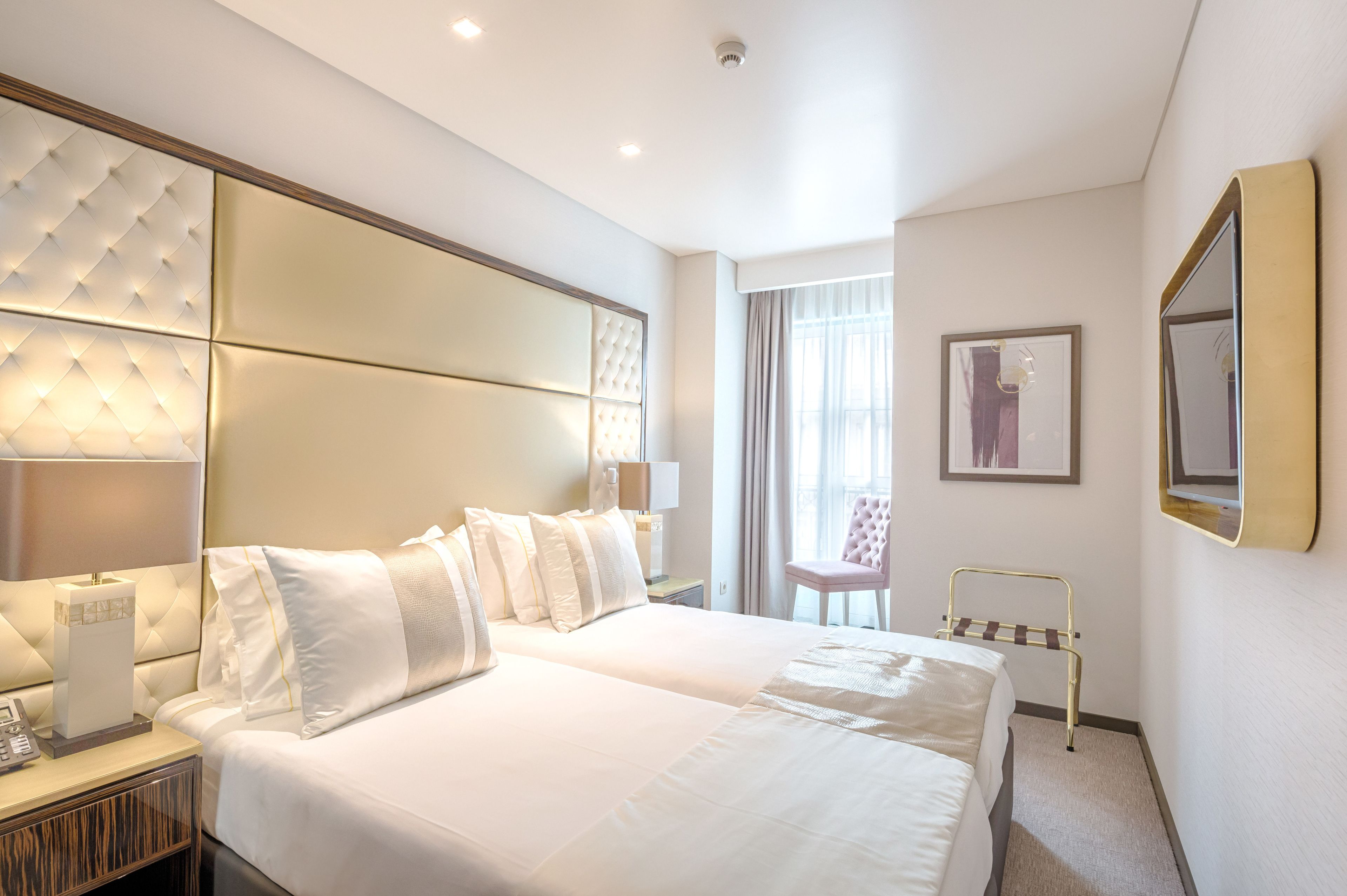 Ett hotellrum med två sängar, en stor, vadderad sänggavel, sänglampor, en telefon vid sängen, en stol och ett litet bord.