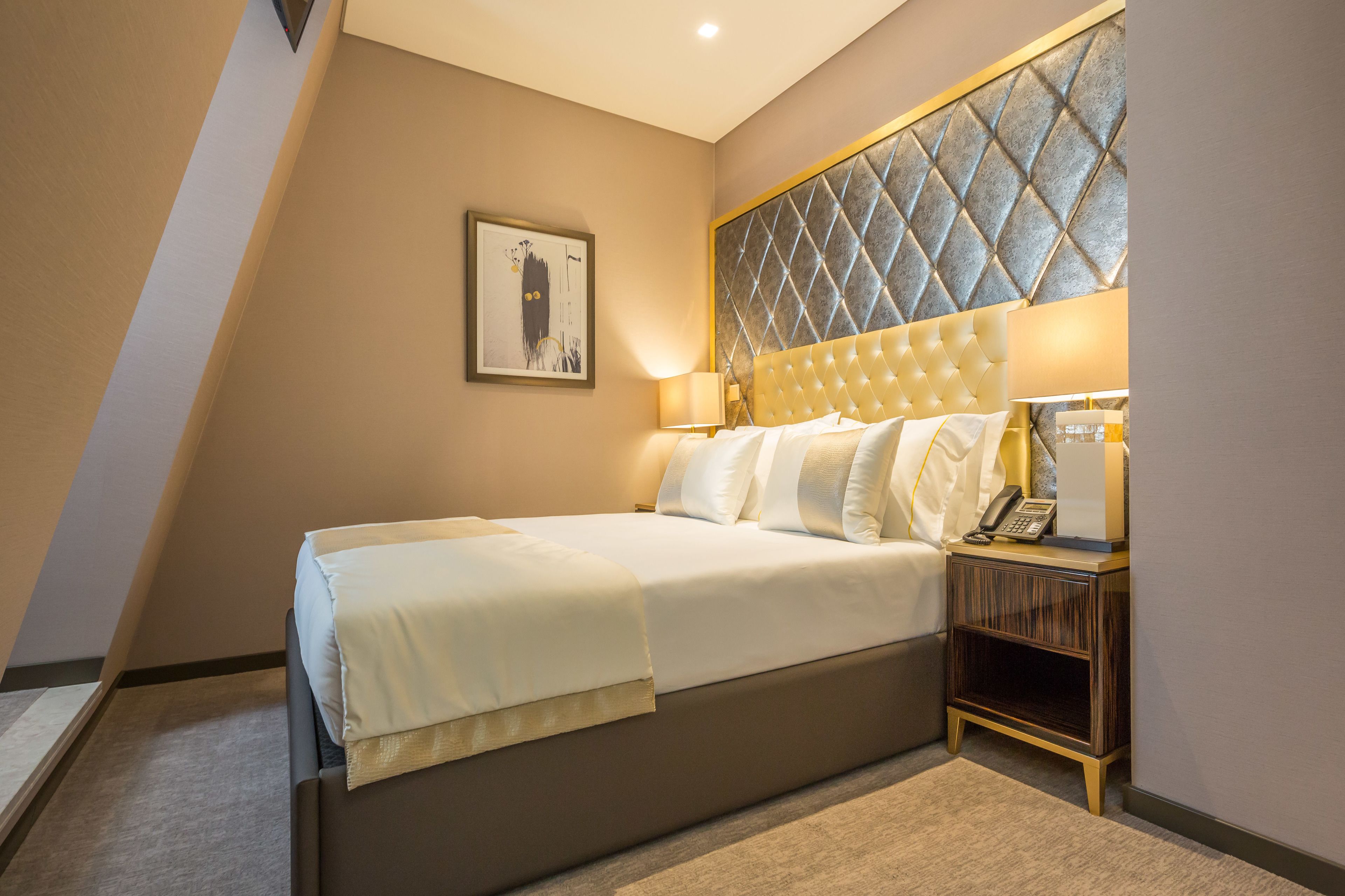 Ein modernes Hotelzimmer mit einem großen Bett, einem Kopfteil mit Rautenmuster, einem Nachttisch mit Lampe und einem gerahmten Kunstwerk an der Wand.