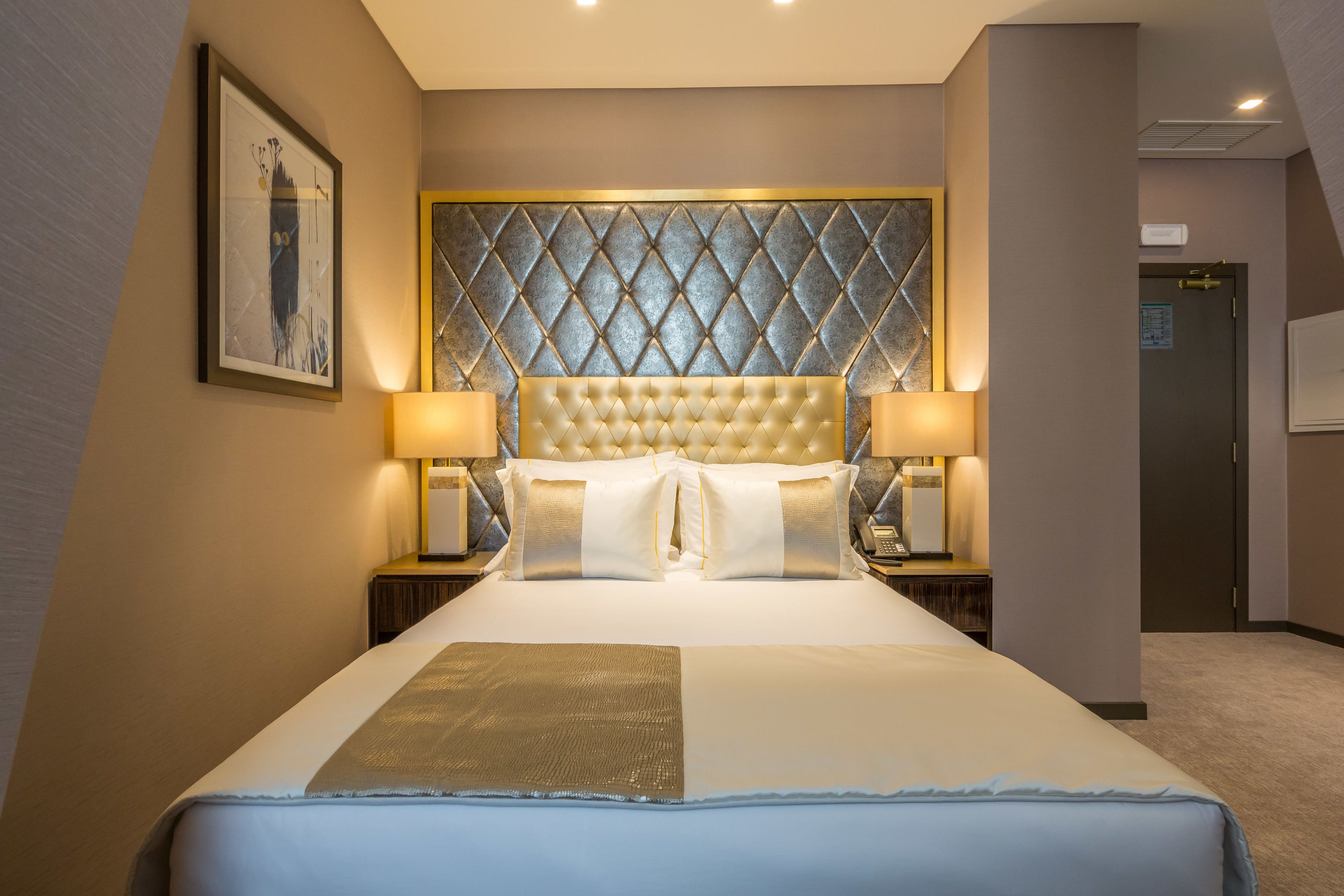 Een hotelkamer met een groot bed, twee nachtkastjes met lampen, een decoratief hoofdeinde en een ingelijst schilderij aan de muur.