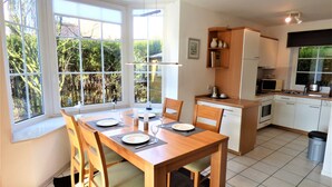A modern kitchen with white cabinets, a dining area with a wooden table and chairs, and large windows offering a view of greenery. - Newly furnished holiday home near the dike well suited for families with WiFi (Dornum-Neßmersiel)