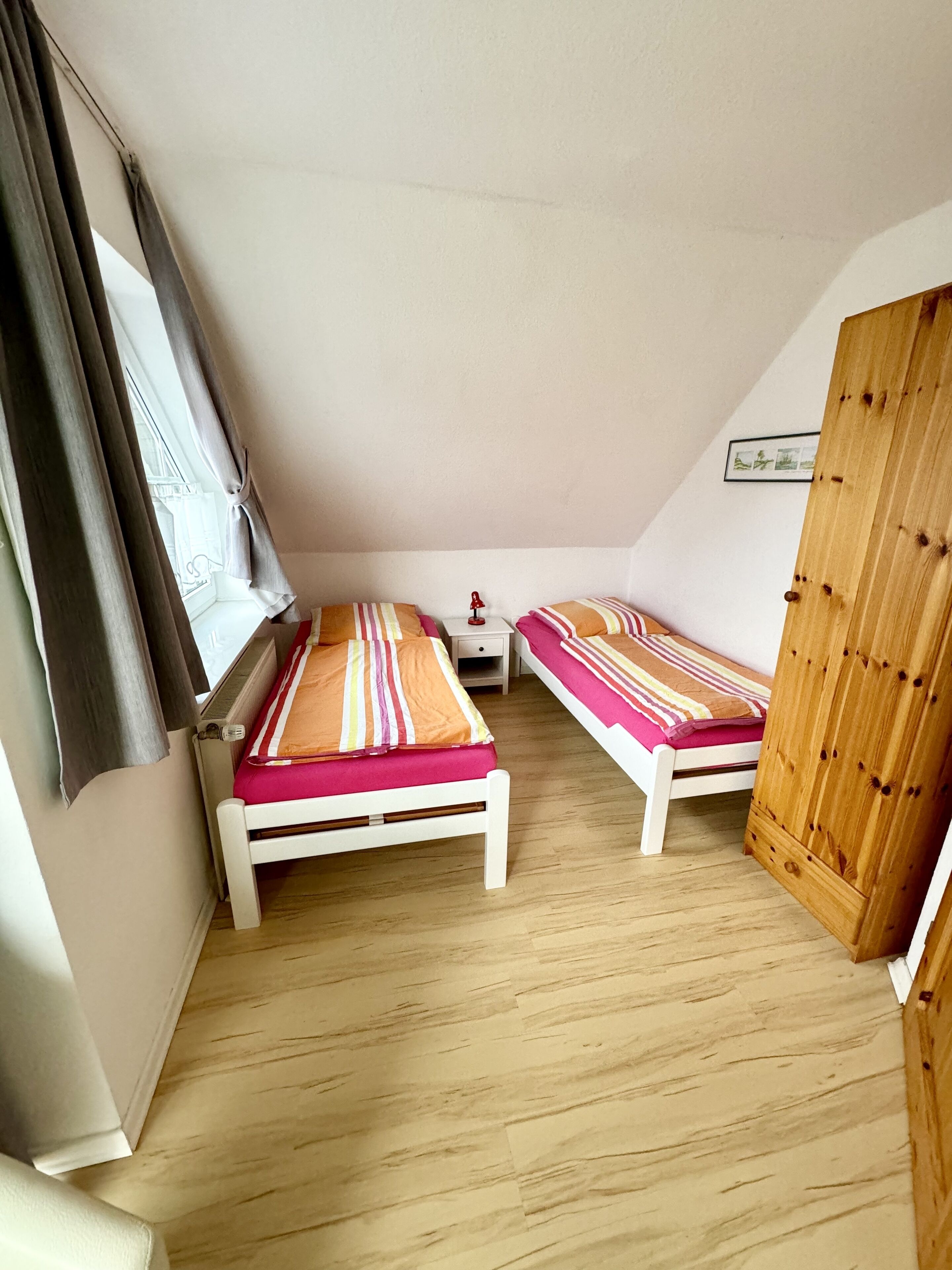 Una habitación pequeña y acogedora con dos camas individuales, una mesita de noche, un radiador y un armario de madera.