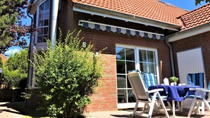A house with a red-tiled roof, a brick exterior, and a large window. There is a patio with outdoor furniture and a small tree. - Newly furnished holiday home near the dike well suited for families with WiFi (Dornum-Neßmersiel)