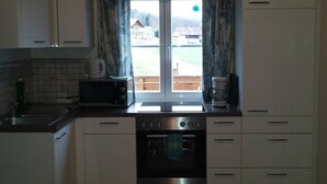 Una cocina con armarios blancos, microondas, horno y ventana con vista a una casa y vegetación.