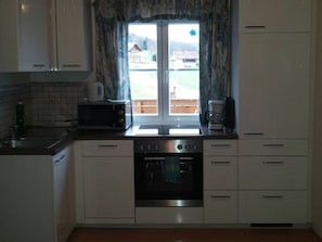 Una cocina con armarios blancos, microondas, horno y ventana con vista a una casa y vegetación.