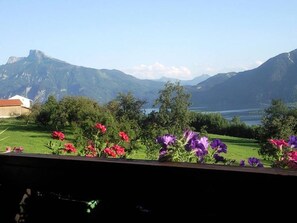 Vista de un paisaje montañoso con un lago, flores en primer plano y un edificio a la izquierda.
