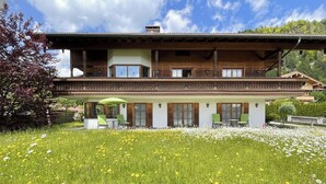A two-story wooden chalet with a balcony, surrounded by a meadow with wildflowers. - (2) FeWo Bergveilchen, Zwei-Raum-Wohnung, ca. 43 qm, mit Terrasse (Reit im Winkl)