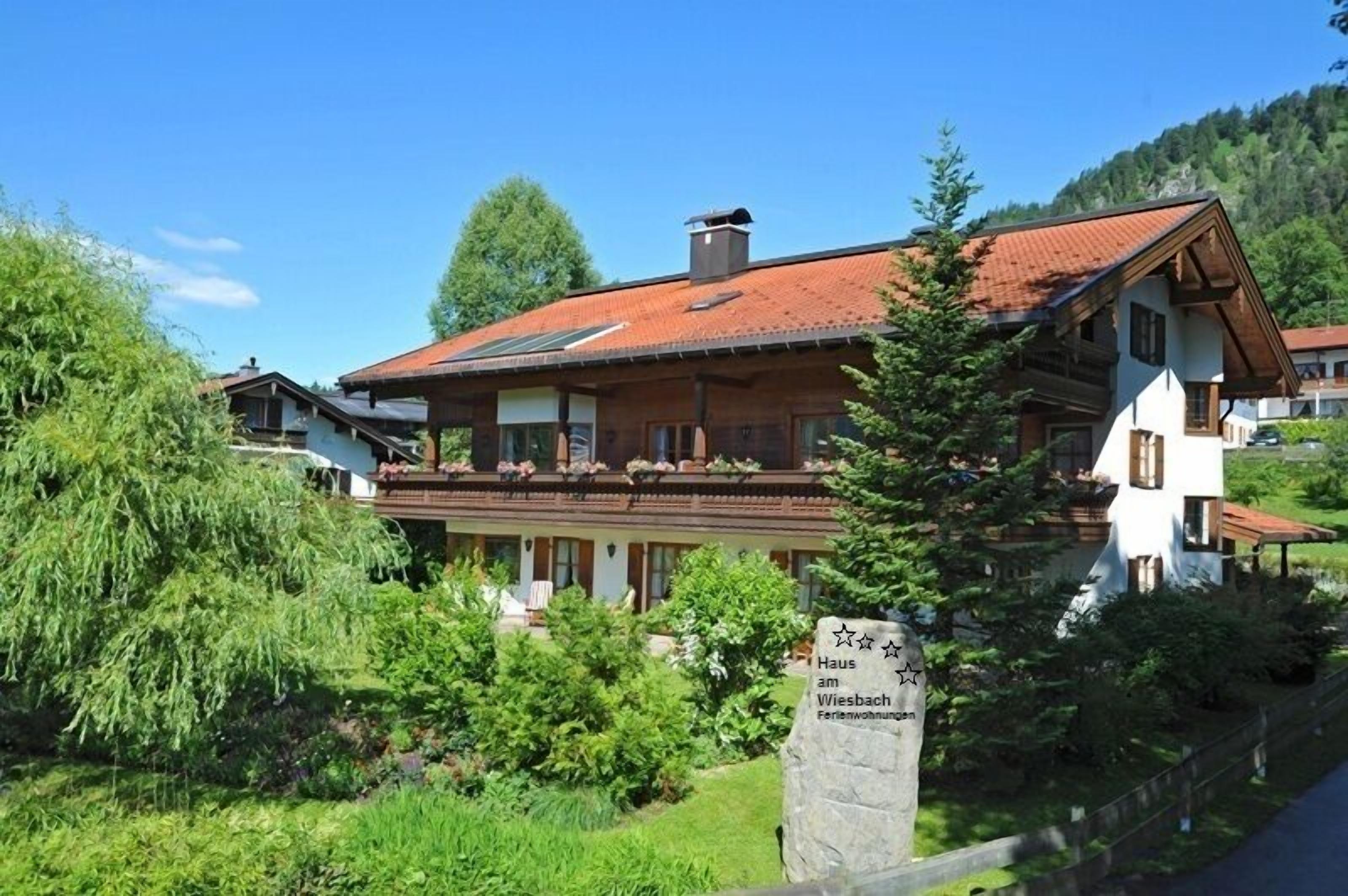 A two-story wooden house with a red-tiled roof, surrounded by lush greenery and a clear blue sky.