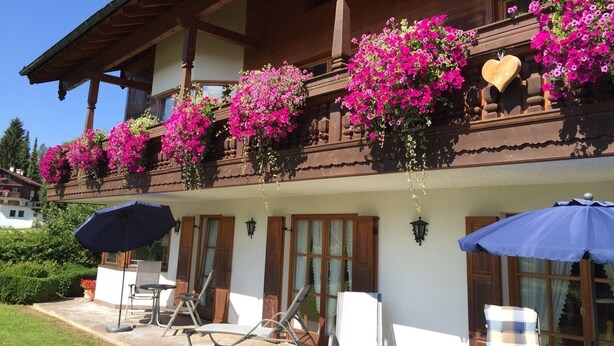 A wooden building with balconies adorned with pink flowers, outdoor seating, and a clear sky. - (2) FeWo Bergveilchen, Zwei-Raum-Wohnung, ca. 43 qm, mit Terrasse (Reit im Winkl)