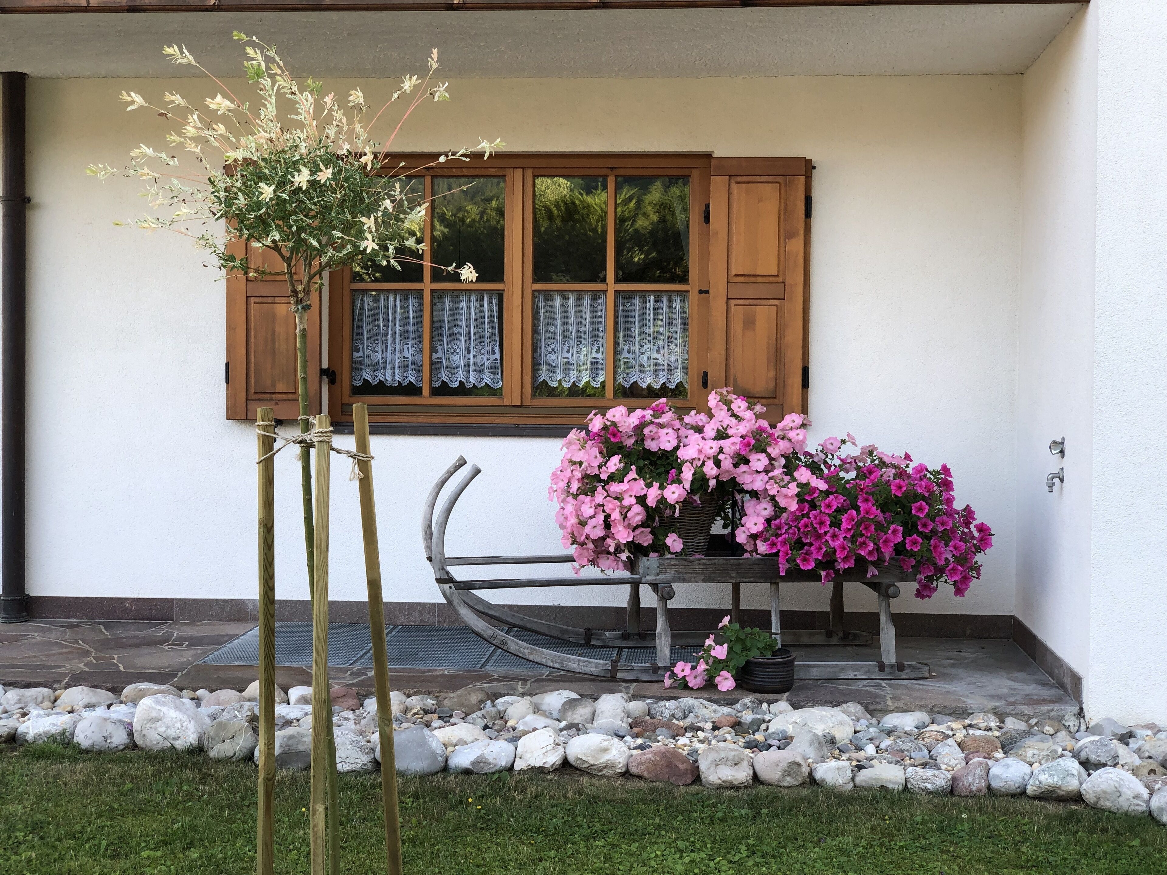 Una casa con puerta de madera, ventana con cortinas de encaje y un jardín con flores rosadas y un banco de madera.