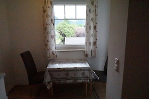 A small room with a table and a chair, a window with curtains, and a view of trees and buildings outside. - Apartment Sonntagshorn 36sqm, balcony, 1 bedroom, living room, WLAN, 3 p. (Ruhpolding)