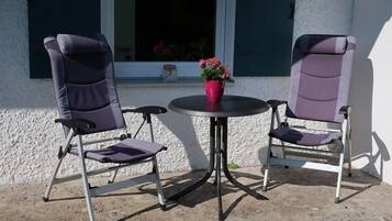 Zwei klappbare Terrassenstühle mit Kissen, ein kleiner runder Tisch und eine rote Blumenvase.