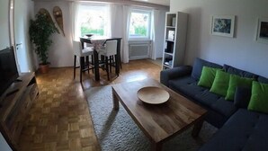 A living room with a wooden coffee table, a dark sofa with green cushions, a dining area with a table and chairs, a bookshelf, and a potted plant. - Vacation apartment Thann (Brannenburg)