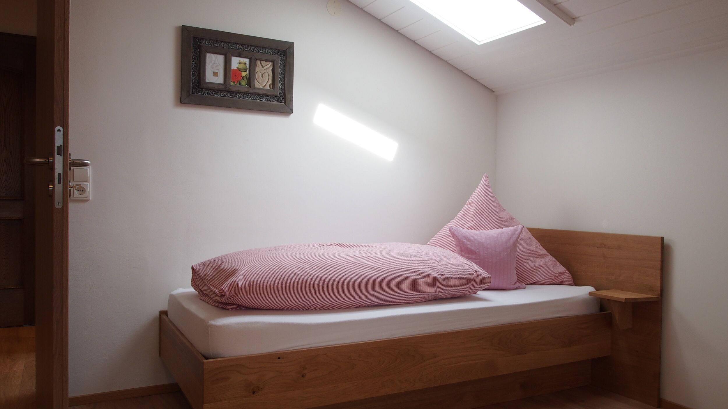 A small, single-bed room with a sloped ceiling and a skylight, featuring a wooden headboard and pink striped bedding.