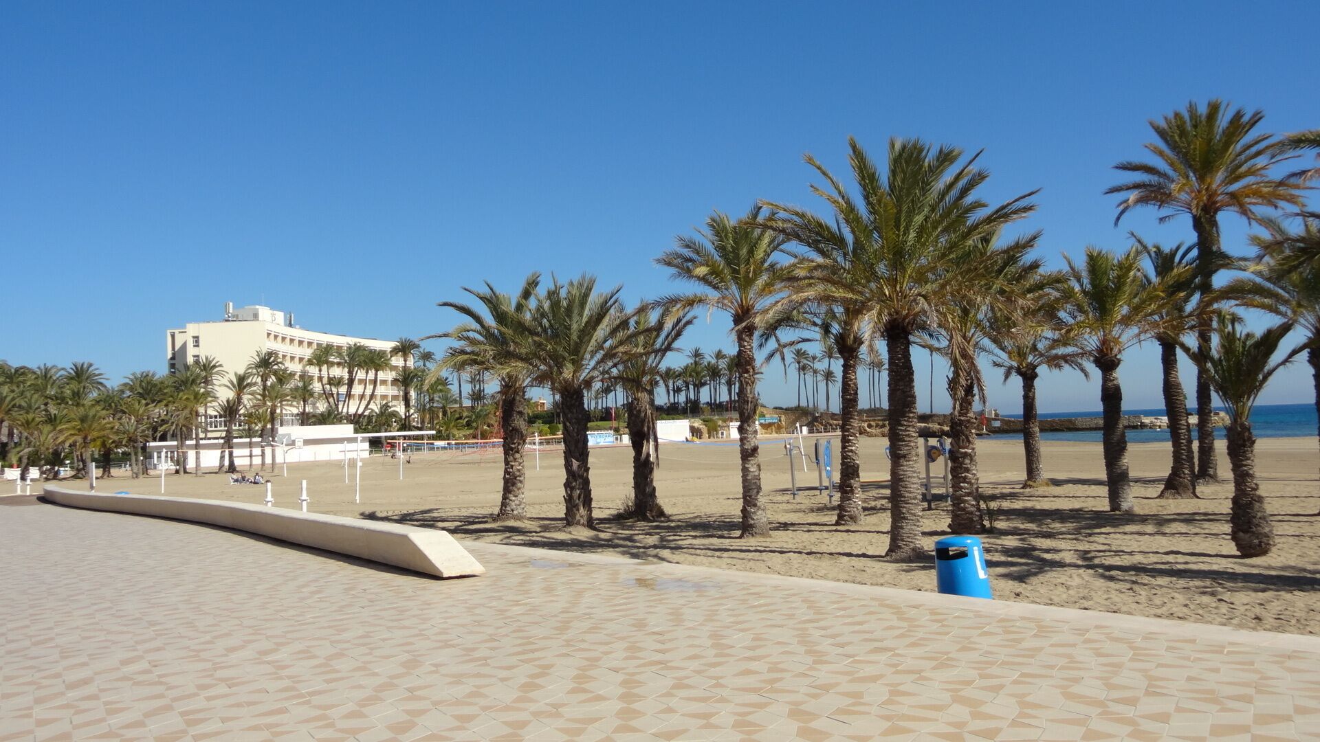 Een strandboulevard met palmbomen, een volleybalveld en een gebouw op de achtergrond.