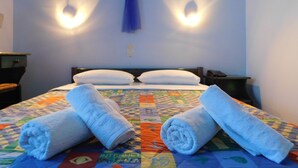 A hotel room with a bed, two pillows, a blue towel, a hat on the wall, and a quilt with an 'INTERNET' design. - Studio Elisabeth (Tinos)