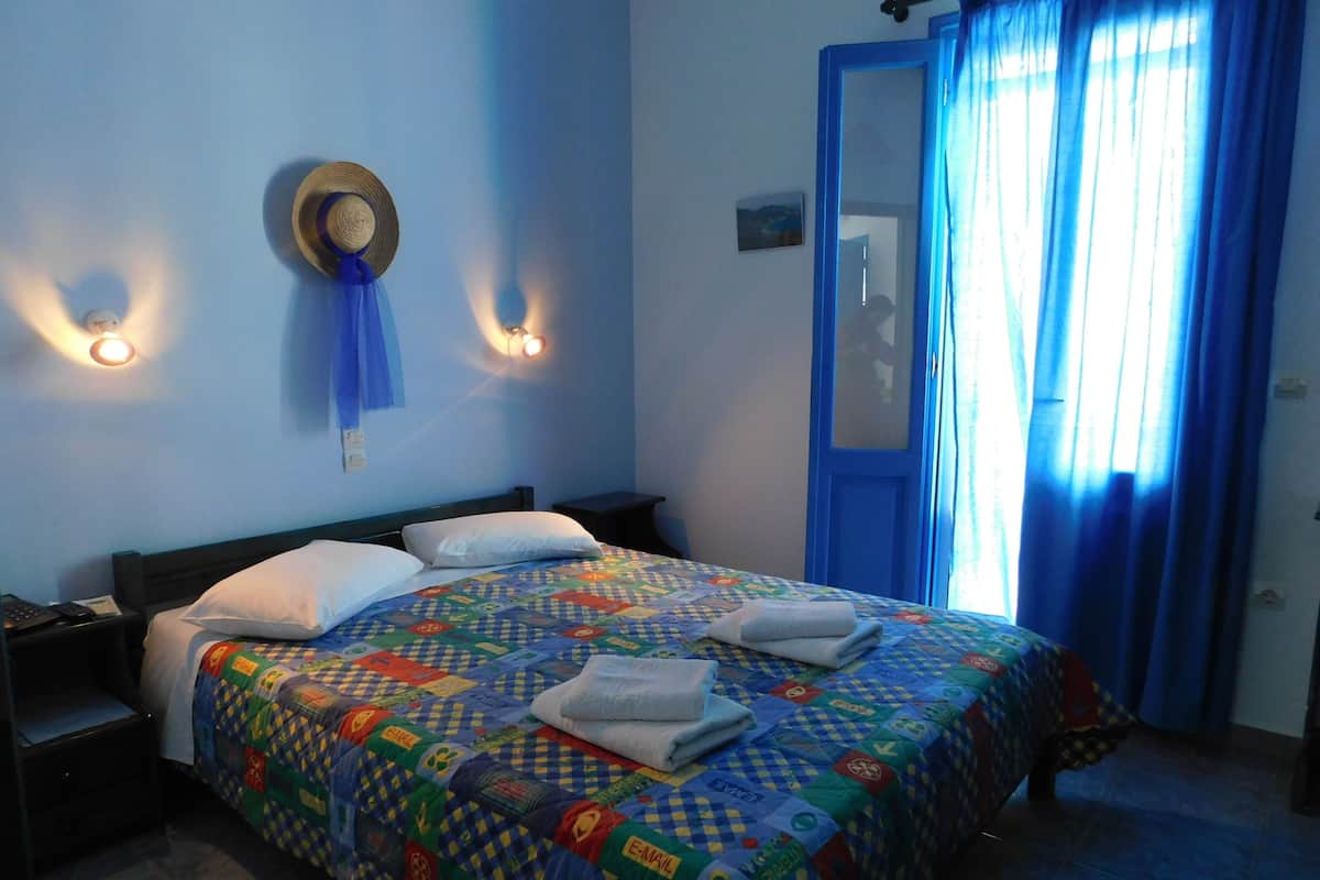 A hotel room with a bed, two bedside tables, a nightstand, a wall-mounted hat, two wall sconces, a door with a blue frame, and a window with blue curtains.