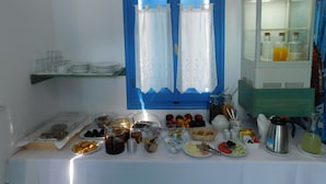 A breakfast buffet with various food items and beverages, a blue-framed window, and a refrigerator with drinks. - Studio Elisabeth (Tinos)