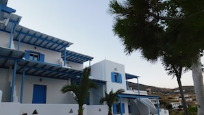 A white building with blue accents, a patio area with seating, and a view of the surrounding landscape. - Studio Elisabeth (Tinos)