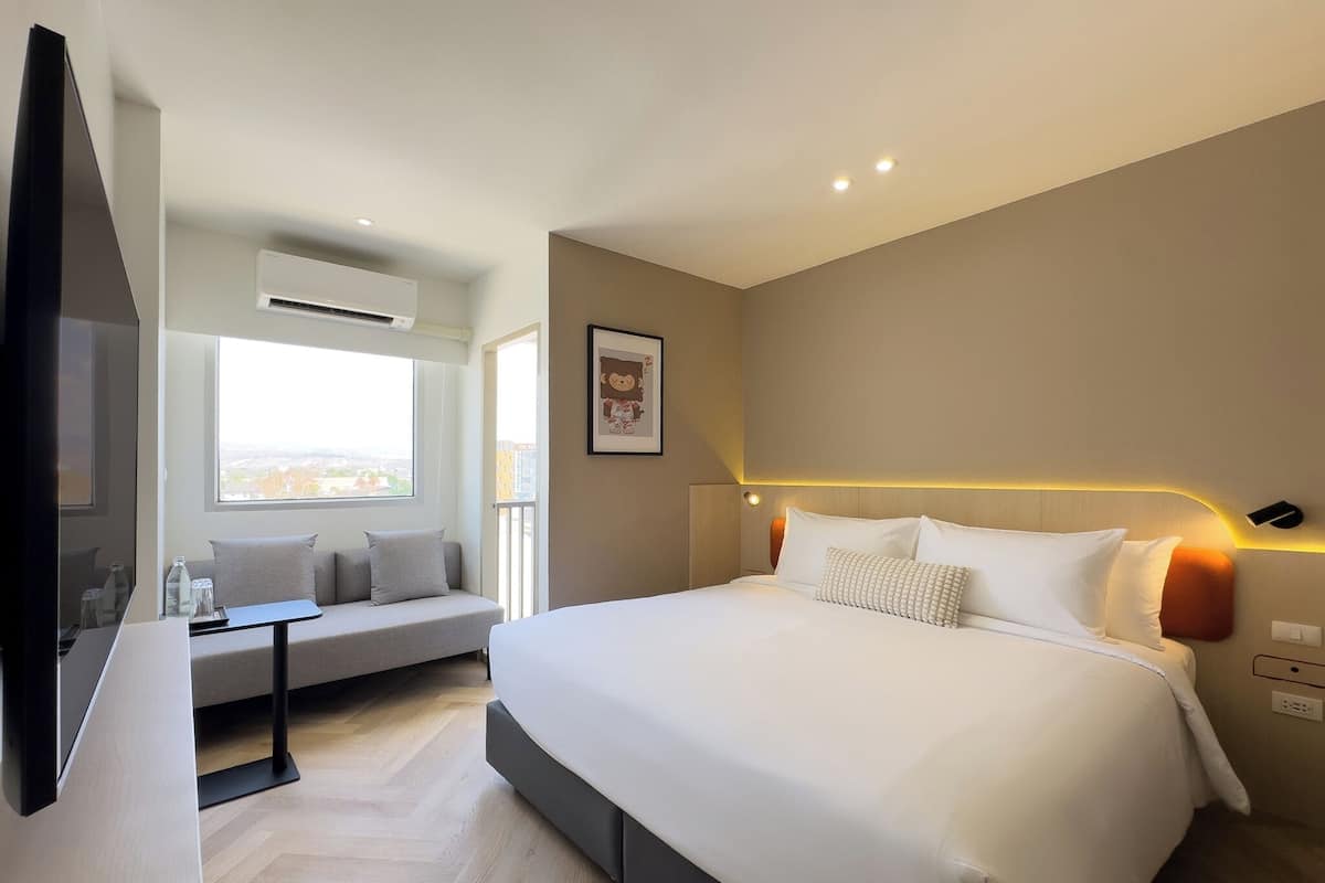 Une chambre d’hôtel moderne dotée d’un grand lit, d’un canapé, d’une télévision et d’une fenêtre donnant sur la ville.