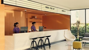Ein moderner Empfangsbereich mit Theke, zwei Mitarbeitern, einer Rezeption und einer Sitzecke mit Sessel und Hocker.