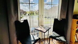 A balcony with two black chairs and a small table, large windows with a view of houses and mountains, and a bookshelf with books. - Vacation home in Neßmersiel, quiet cul-de-sac location, close to the dyke (Dornum-Neßmersiel)