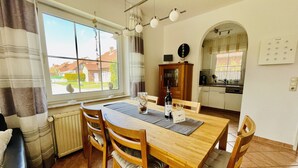 A dining area with a wooden table and chairs, a window with curtains, a kitchen in the background, and a clock on the wall. - Vacation home in Neßmersiel, quiet cul-de-sac location, close to the dyke (Dornum-Neßmersiel)
