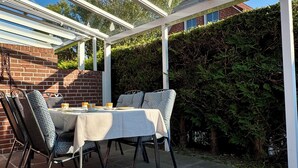 An outdoor patio with a glass enclosure, a table set for dining, and a brick wall in the background. - Vacation home in Neßmersiel, quiet cul-de-sac location, close to the dyke (Dornum-Neßmersiel)