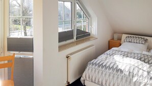 Una camera da letto con soffitto spiovente, una finestra con persiane, un termosifone, un comodino in legno, un letto con biancheria a motivi decorativi e una sedia.