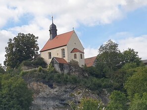 A church with a red roof, a bell tower, and a cross on top, surrounded by greenery and trees.