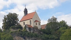 Eine Kirche mit rotem Dach, einem Glockenturm und einem Kreuz auf der Spitze, umgeben von Grünflächen und Bäumen.